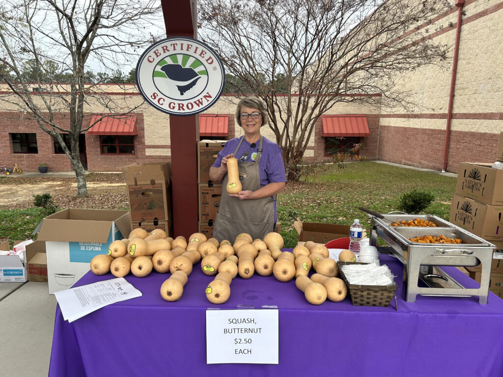 A middle aged woman stands outdoors behind a table holding up a butternut squash. Many butternut squash are laid out on the table, as well as warming trays with roasted butternut squash samples. The Certified SC Grown oval logo sign hangs on a vertical beam behind her.